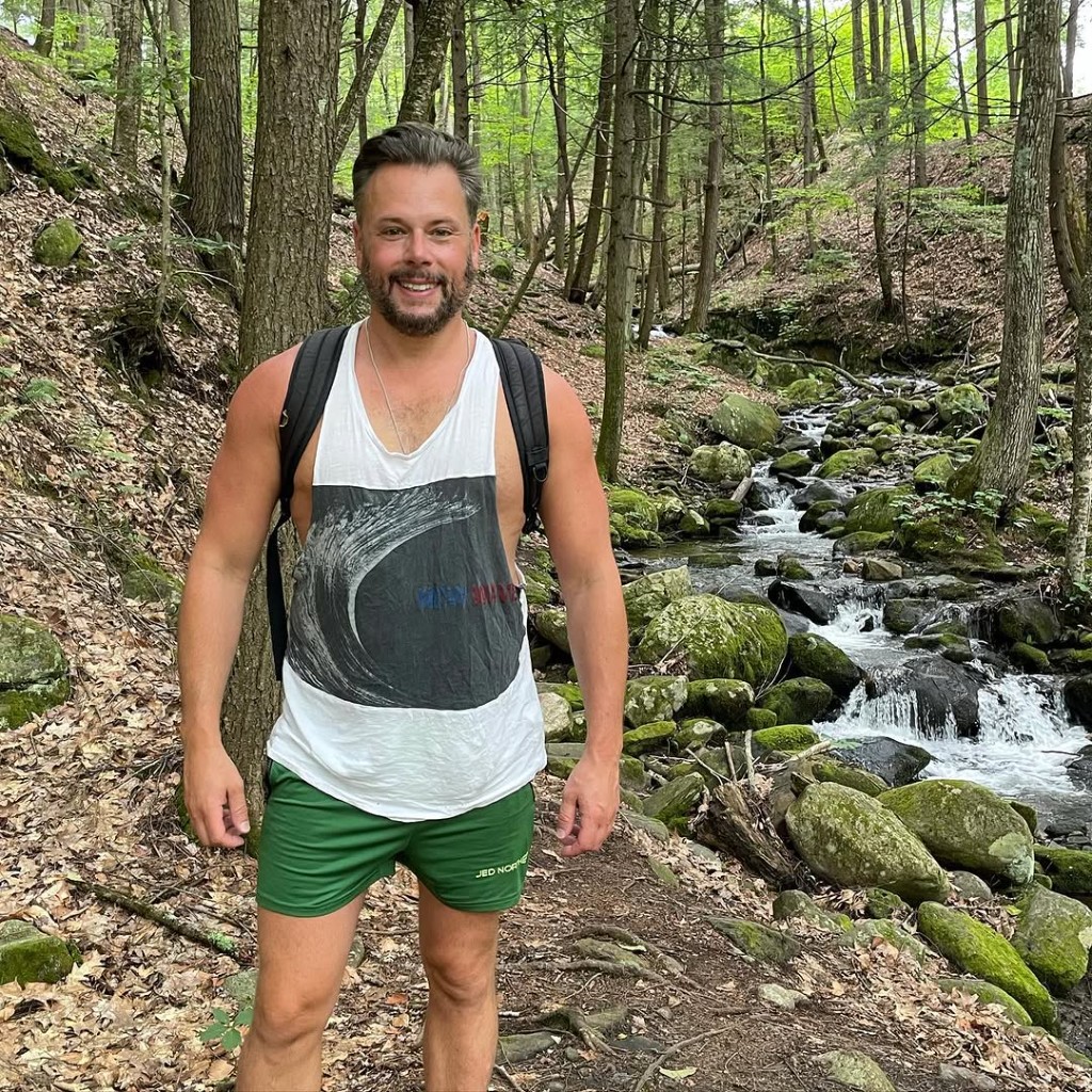 Rock River — hiker by a side stream A smiling hiker with a backpack stands on a mossy wooded trail beside a small rocky stream at Rock River, Vermont — bright summer greens
